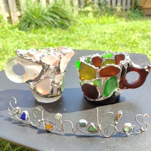 May include: Two small cups made of sea glass, one is pink and the other is green, brown, and yellow. The cups are sitting on a black surface with a wire sculpture that says "Sea Stained Glass".