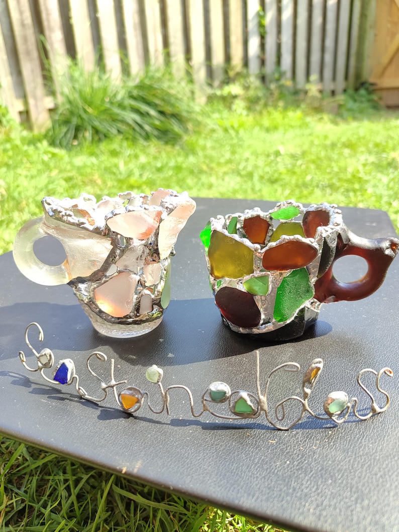 May include: Two small cups made of sea glass, one is pink and the other is green, brown, and yellow. The cups are sitting on a black surface with a wire sculpture that says "Sea Stained Glass".