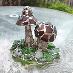 May include: A handcrafted glass art piece featuring two mushroom sculptures. The mushroom caps are made of brown and amber glass pieces, while the stems are white and silver. The base is composed of green glass pieces, all held together with silver solder.