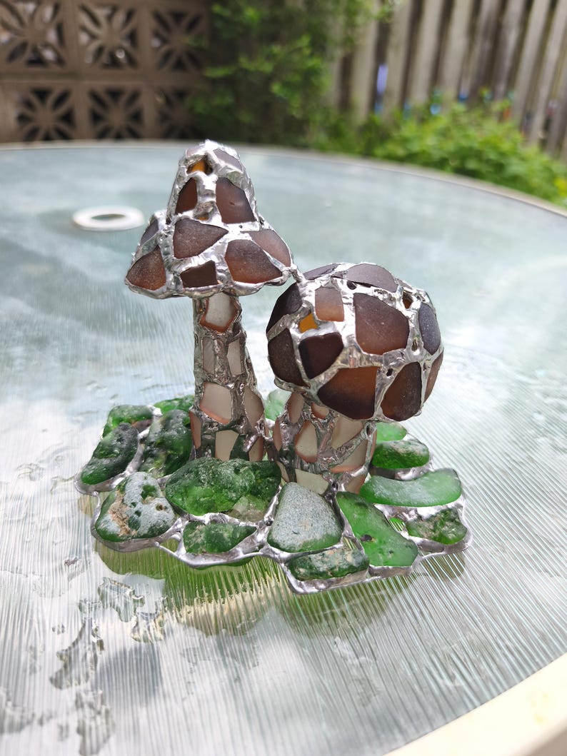 May include: A handcrafted glass art piece featuring two mushroom sculptures. The mushroom caps are made of brown and amber glass pieces, while the stems are white and silver. The base is composed of green glass pieces, all held together with silver solder.