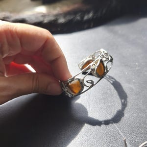 May include: A silver-toned cuff bracelet with amber-colored, teardrop-shaped stones. The bracelet has an open design with decorative filigree and is held by a hand against a dark background. The stones are a warm, golden hue.