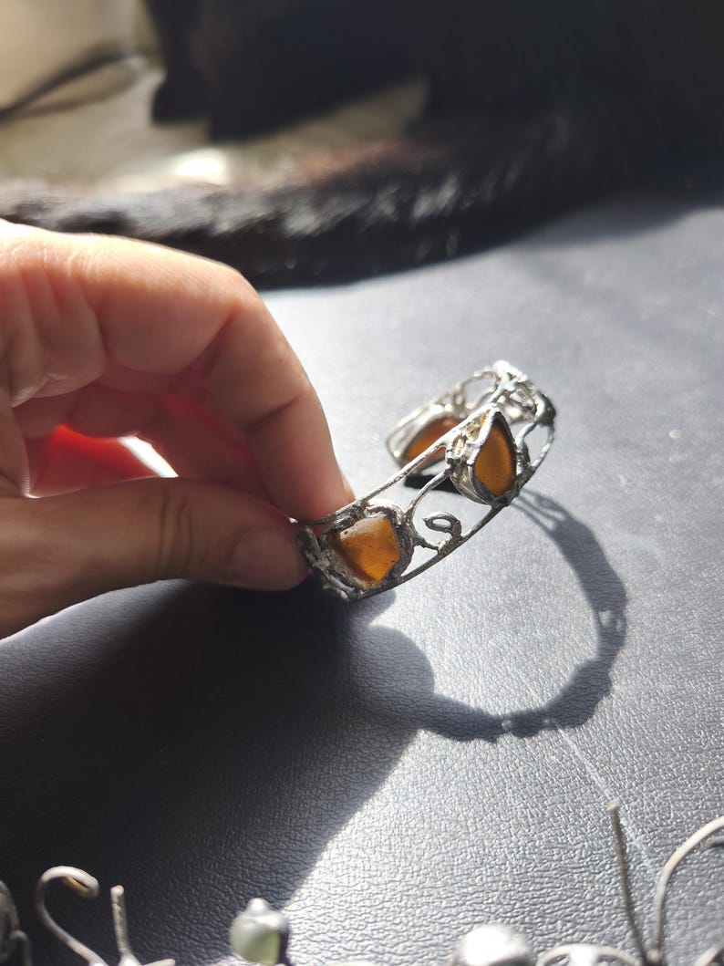 May include: A silver-toned cuff bracelet with amber-colored, teardrop-shaped stones. The bracelet has an open design with decorative filigree and is held by a hand against a dark background. The stones are a warm, golden hue.