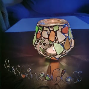 May include: A glass candle holder with a mosaic design in shades of green, orange, and white. The holder is lit with a small white candle.