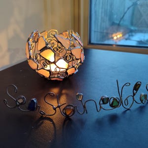 May include: A glass candle holder with a round shape and a mosaic design. The holder is made of clear glass with pink and white pieces. The candle holder is lit and casting a warm glow. The word "Sea Stones" is spelled out in wire with glass beads.