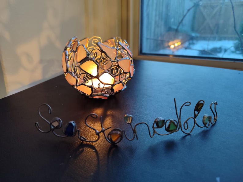 May include: A glass candle holder with a round shape and a mosaic design. The holder is made of clear glass with pink and white pieces. The candle holder is lit and casting a warm glow. The word "Sea Stones" is spelled out in wire with glass beads.