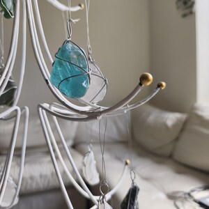 May include: A white metal mobile with various sea glass pieces in shades of blue, green, and clear. Each piece is wrapped in wire and suspended from the mobile's curved arms. The mobile has a decorative, artistic design.