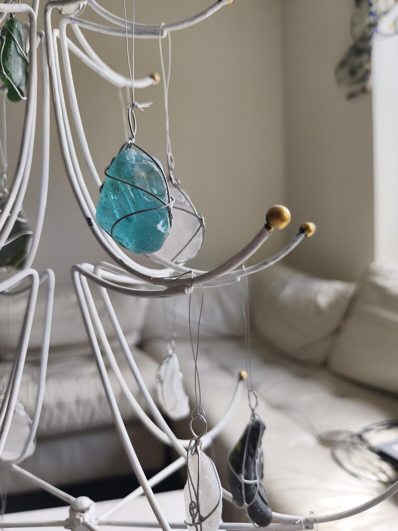 May include: A white metal mobile with various sea glass pieces in shades of blue, green, and clear. Each piece is wrapped in wire and suspended from the mobile's curved arms. The mobile has a decorative, artistic design.