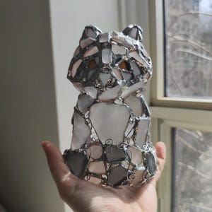 May include: A handmade stained glass cat sculpture. The cat is made from clear, white, grey, and amber glass pieces, joined with silver-coloured metal. The sculpture is held in a person's hand. The cat is a decorative item.