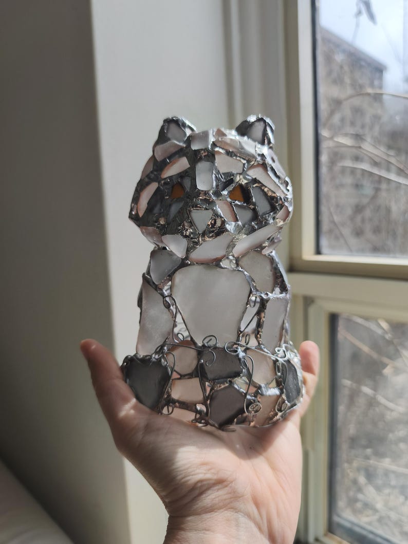 May include: A handmade stained glass cat sculpture. The cat is made from clear, white, grey, and amber glass pieces, joined with silver-coloured metal. The sculpture is held in a person's hand. The cat is a decorative item.