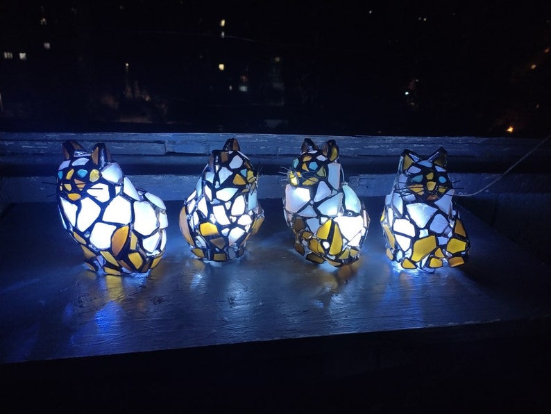 May include: Four stained glass cat figurines with white, yellow, and black glass pieces. The cats are illuminated from within, casting a warm glow.