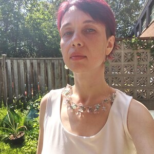 May include: A woman wearing a delicate necklace with clear and pastel-colored floral accents. The necklace is set against a white top. The background includes a wooden fence, greenery, and a sunny outdoor setting.