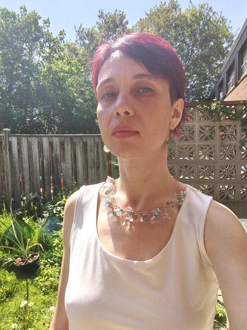 May include: A woman wearing a delicate necklace with clear and pastel-colored floral accents. The necklace is set against a white top. The background includes a wooden fence, greenery, and a sunny outdoor setting.