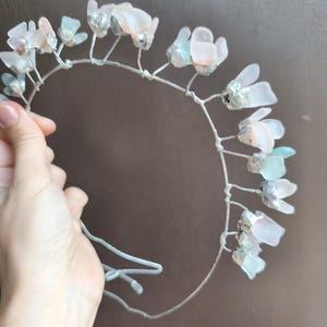 May include: A delicate floral crown featuring translucent, petal-shaped accents in shades of pink, blue, and white. The flowers are attached to a silver-colored wire frame. The crown is held by a hand against a brown background. Perfect for weddings or special occasions.