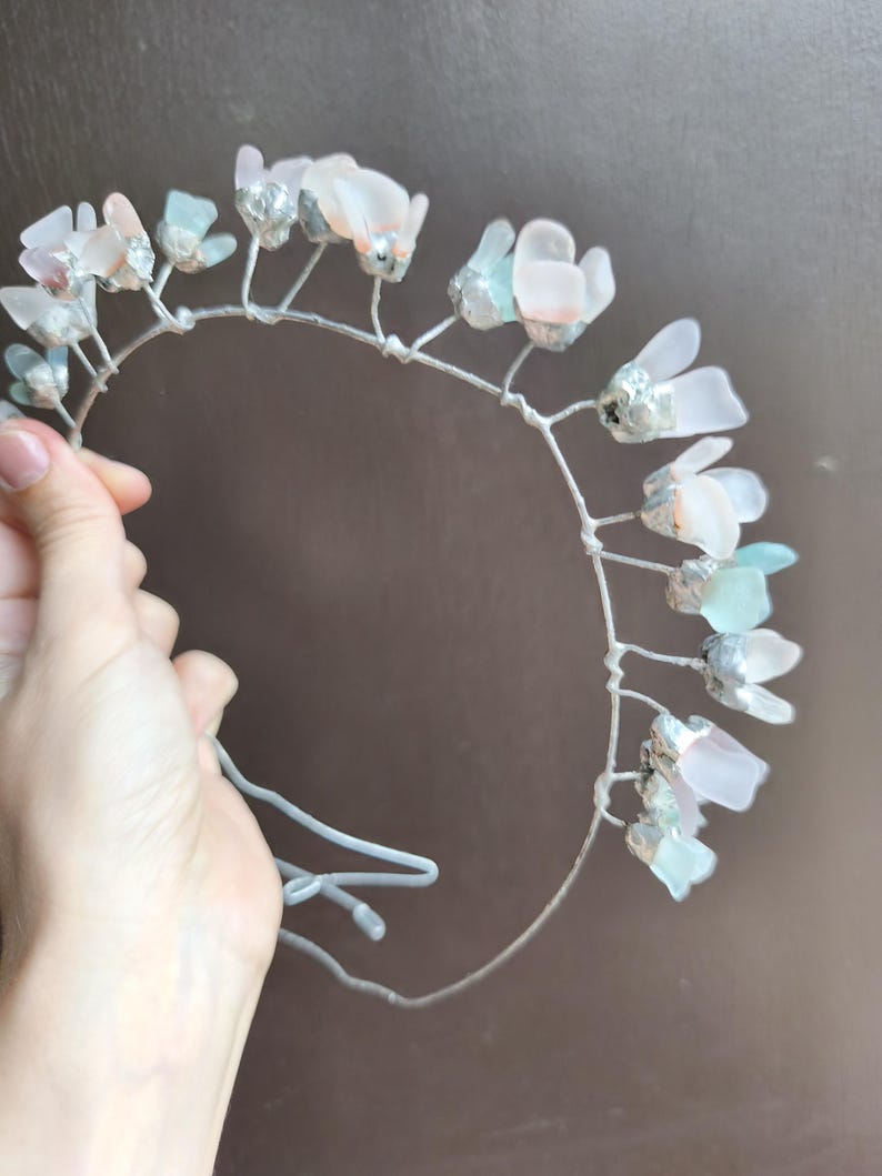 May include: A delicate floral crown featuring translucent, petal-shaped accents in shades of pink, blue, and white. The flowers are attached to a silver-colored wire frame. The crown is held by a hand against a brown background. Perfect for weddings or special occasions.