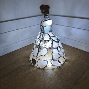 以下が含まれることがあります： 暗い金属製のワイヤーで繋ぎ合わされた白と水色のシーグラスの破片で構成された、ベル型の広いドレスを着た人物の形をした装飾的な置物。内部から小さなLEDストリングライトで照らされています。
