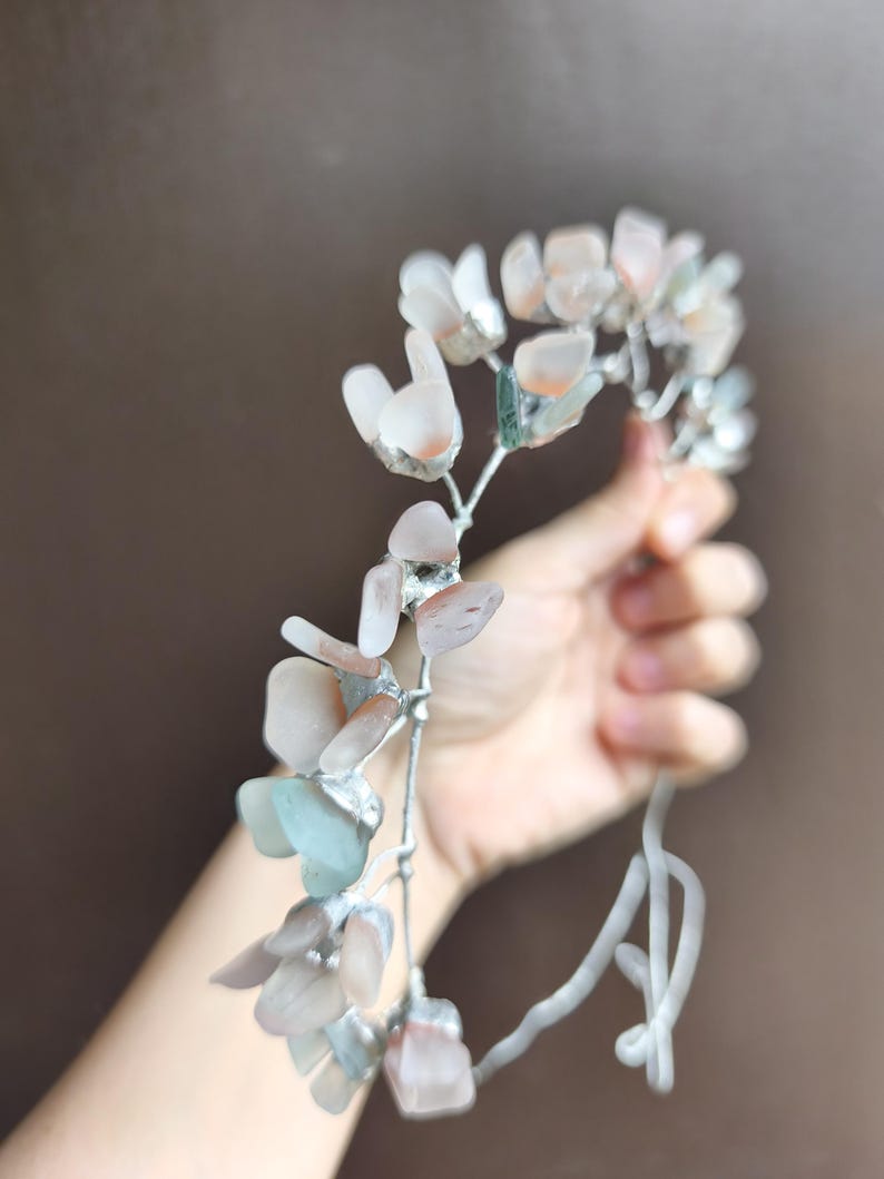May include: A delicate floral headpiece featuring pale pink, peach, and blue-green gemstone petals. The gemstones are set on a silver-colored wire frame, creating a whimsical and ethereal design. Perfect for weddings or special occasions.