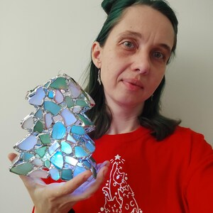K&ouml;nnte beinhalten: Eine Frau mit gr&uuml;nen Haaren h&auml;lt einen Weihnachtsbaumschmuck aus Glasmalerei. Der Schmuck besteht aus kleinen Glassplittern in Blaut&ouml;nen, Gr&uuml;n- und Wei&szlig;t&ouml;nen. Der Schmuck wird von innen beleuchtet und erzeugt so ein warmes Leuchten.