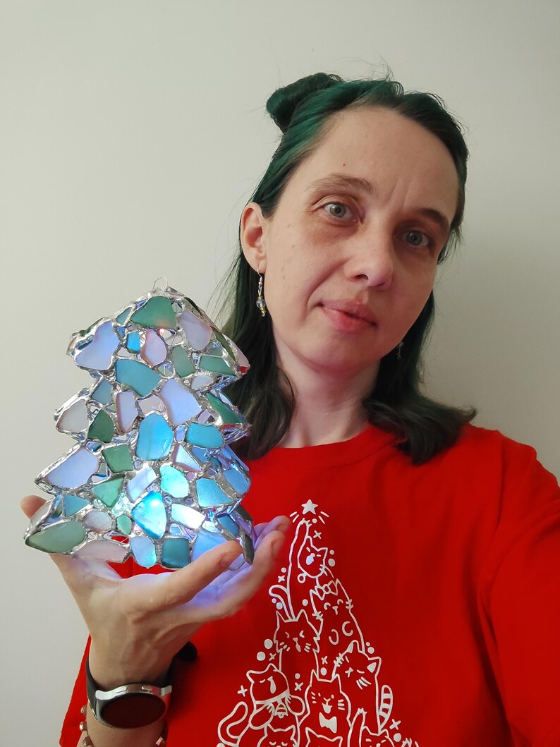 K&ouml;nnte beinhalten: Eine Frau mit gr&uuml;nen Haaren h&auml;lt einen Weihnachtsbaumschmuck aus Glasmalerei. Der Schmuck besteht aus kleinen Glassplittern in Blaut&ouml;nen, Gr&uuml;n- und Wei&szlig;t&ouml;nen. Der Schmuck wird von innen beleuchtet und erzeugt so ein warmes Leuchten.