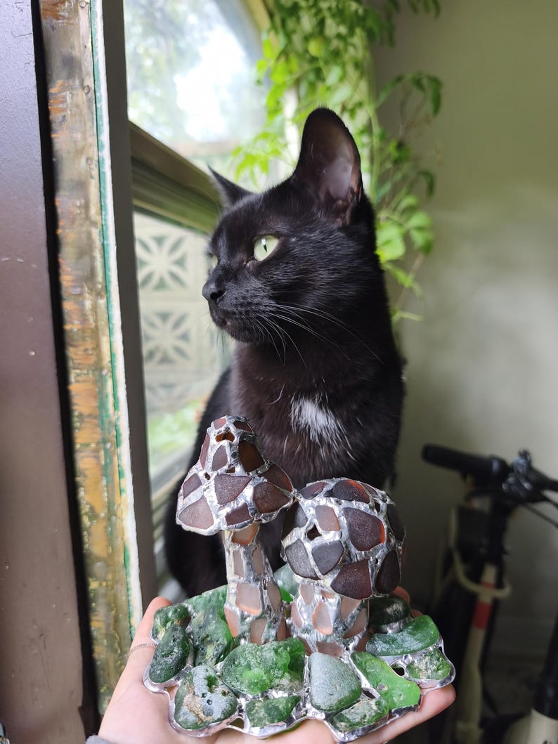 May include: A black cat with green eyes looks out a window. In front of the cat is a decorative mushroom sculpture made of brown and clear glass pieces, set on a base of green glass.
