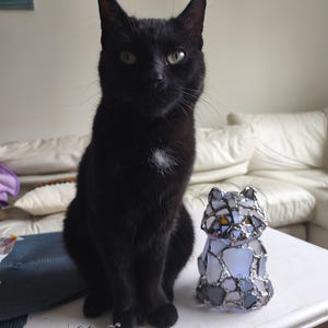 May include: A black cat with green eyes sits beside a stained glass cat ornament. The ornament is made of blue, grey, and clear glass pieces, held together with a silver metal frame. The cat has a small white patch on its chest. The cat is on a white table.