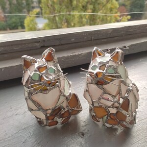 May include: Two stained glass cat figurines, one white with brown and green accents, the other brown with white and green accents. The cats are sitting on a gray surface.