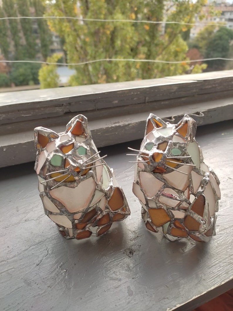 May include: Two stained glass cat figurines, one white with brown and green accents, the other brown with white and green accents. The cats are sitting on a gray surface.