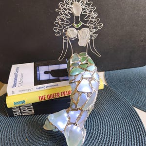 Puede incluir: Una escultura de sirena hecha de alambre plateado y vidrio marino. La figura presenta una cabeza y torso formados por alambre con detalles de vidrio, y una cola construida con piezas de vidrio marino iridiscente, verde y azul unidas por metal.