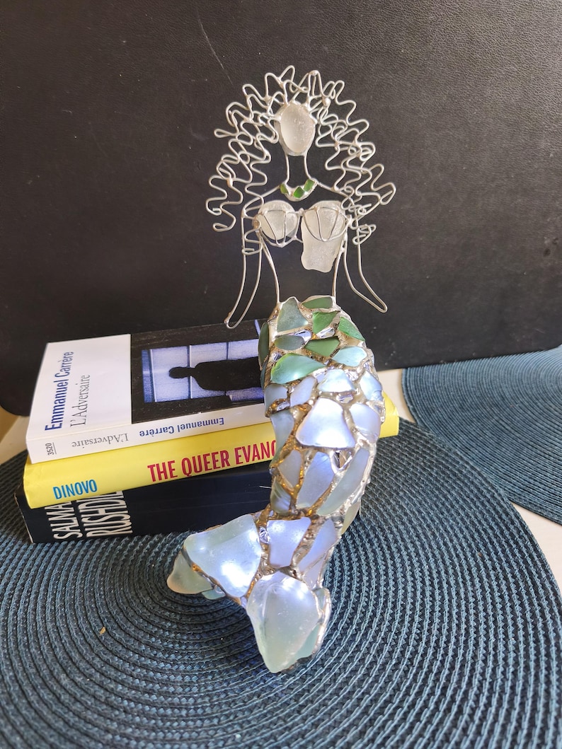 Puede incluir: Una escultura de sirena hecha de alambre plateado y vidrio marino. La figura presenta una cabeza y torso formados por alambre con detalles de vidrio, y una cola construida con piezas de vidrio marino iridiscente, verde y azul unidas por metal.