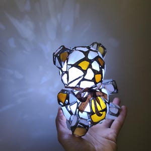 May include: A stained glass teddy bear lamp with a brown ribbon bow. The bear is made of white, yellow, and brown glass pieces, and is lit from within. The lamp is held in a hand.