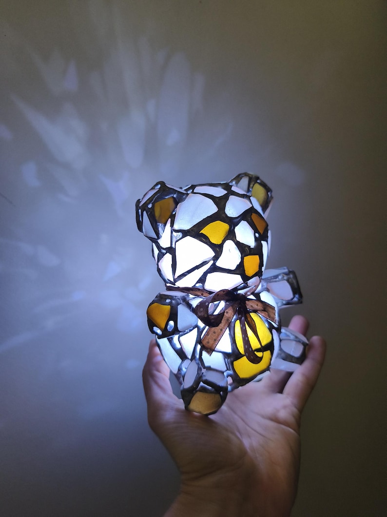 May include: A stained glass teddy bear lamp with a brown ribbon bow. The bear is made of white, yellow, and brown glass pieces, and is lit from within. The lamp is held in a hand.