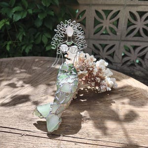 Puede incluir: Una escultura de sirena hecha de alambre y vidrio marino. La figura presenta una cabeza y cabello formados con alambre, un corpi&ntilde;o de vidrio marino blanco y una cola construida con piezas de vidrio marino verde unidas con soldadura de plata. La sirena est&aacute; colocada contra un trozo de coral blanco.