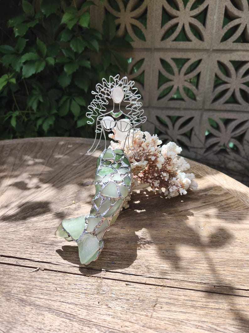 Puede incluir: Una escultura de sirena hecha de alambre y vidrio marino. La figura presenta una cabeza y cabello formados con alambre, un corpi&ntilde;o de vidrio marino blanco y una cola construida con piezas de vidrio marino verde unidas con soldadura de plata. La sirena est&aacute; colocada contra un trozo de coral blanco.