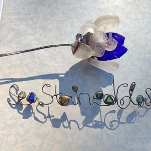 Op de afbeelding: Een draadbeeld van een bloem gemaakt met zeeglas. De bloem is blauw, wit en helder. De draad is gevormd om "Sea Stone Glass" te spellen met extra zeeglasstukken die in de letters zijn verwerkt.