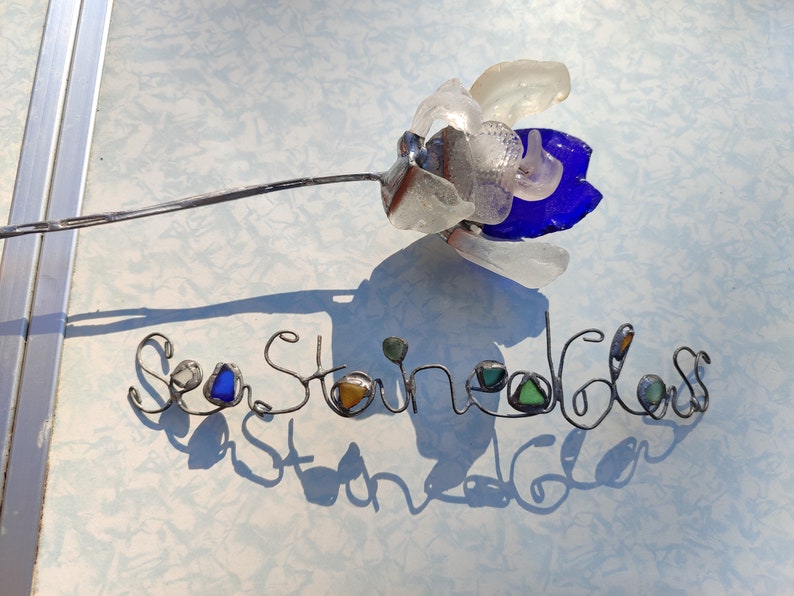Op de afbeelding: Een draadbeeld van een bloem gemaakt met zeeglas. De bloem is blauw, wit en helder. De draad is gevormd om "Sea Stone Glass" te spellen met extra zeeglasstukken die in de letters zijn verwerkt.