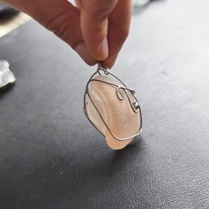 May include: A handcrafted pendant featuring a smooth, peach-colored stone encased in a delicate silver wire frame. The wirework has decorative swirls. The pendant is suspended from a small loop, ready to be worn as a necklace.