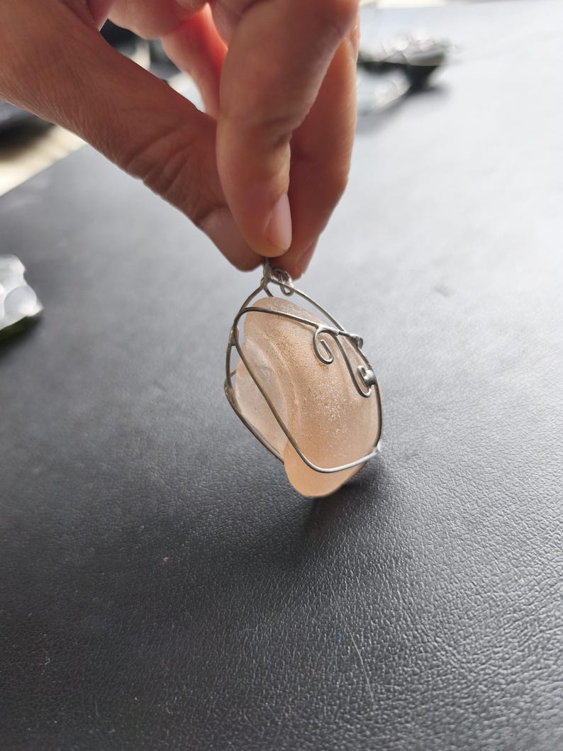 May include: A handcrafted pendant featuring a smooth, peach-colored stone encased in a delicate silver wire frame. The wirework has decorative swirls. The pendant is suspended from a small loop, ready to be worn as a necklace.