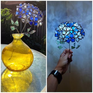 May include: A handcrafted glass hydrangea flower with blue and white petals, green leaves, and a yellow glass vase. The flower is made of individual glass pieces, creating a mosaic effect. The vase is round and has a wide base.