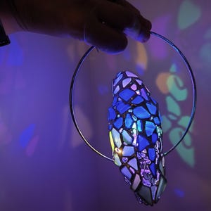 May include: A stained glass pendant lamp with a blue and purple mosaic design. The lamp is suspended from a silver metal ring. The lamp is illuminated, casting colorful light patterns on the background.