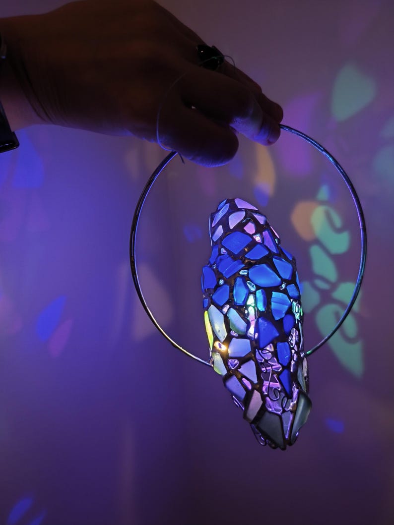May include: A stained glass pendant lamp with a blue and purple mosaic design. The lamp is suspended from a silver metal ring. The lamp is illuminated, casting colorful light patterns on the background.