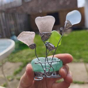 May include: A handcrafted glass flower sculpture. The sculpture features sea glass flowers in shades of pink, blue, and green, set in a teal sea glass base. The flowers are connected with silver wire. A unique decorative item.