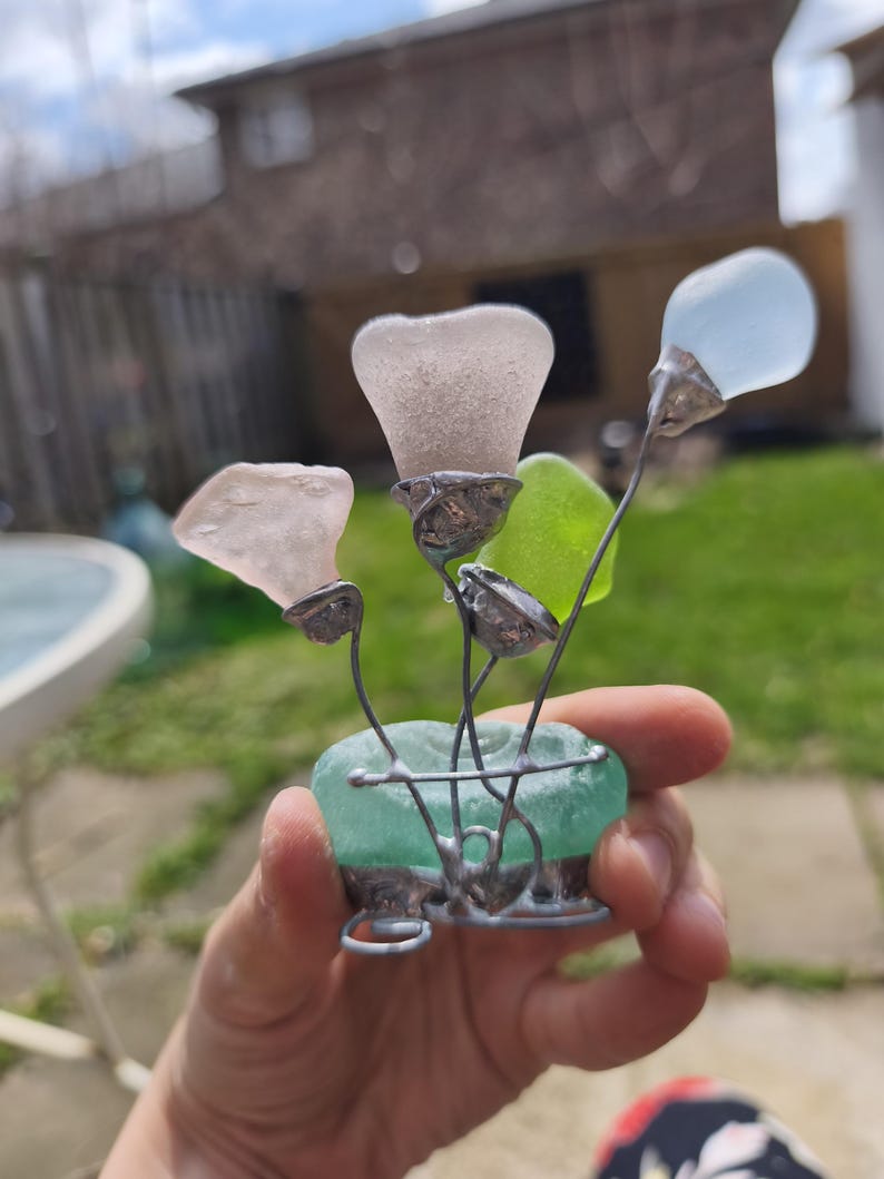 May include: A handcrafted glass flower sculpture. The sculpture features sea glass flowers in shades of pink, blue, and green, set in a teal sea glass base. The flowers are connected with silver wire. A unique decorative item.