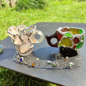 May include: Two small cups made of sea glass and wire. The cups are in the shape of a traditional teacup and are sitting on a black surface. The word "Sea Glass" is spelled out in wire with sea glass pieces used as the letters.