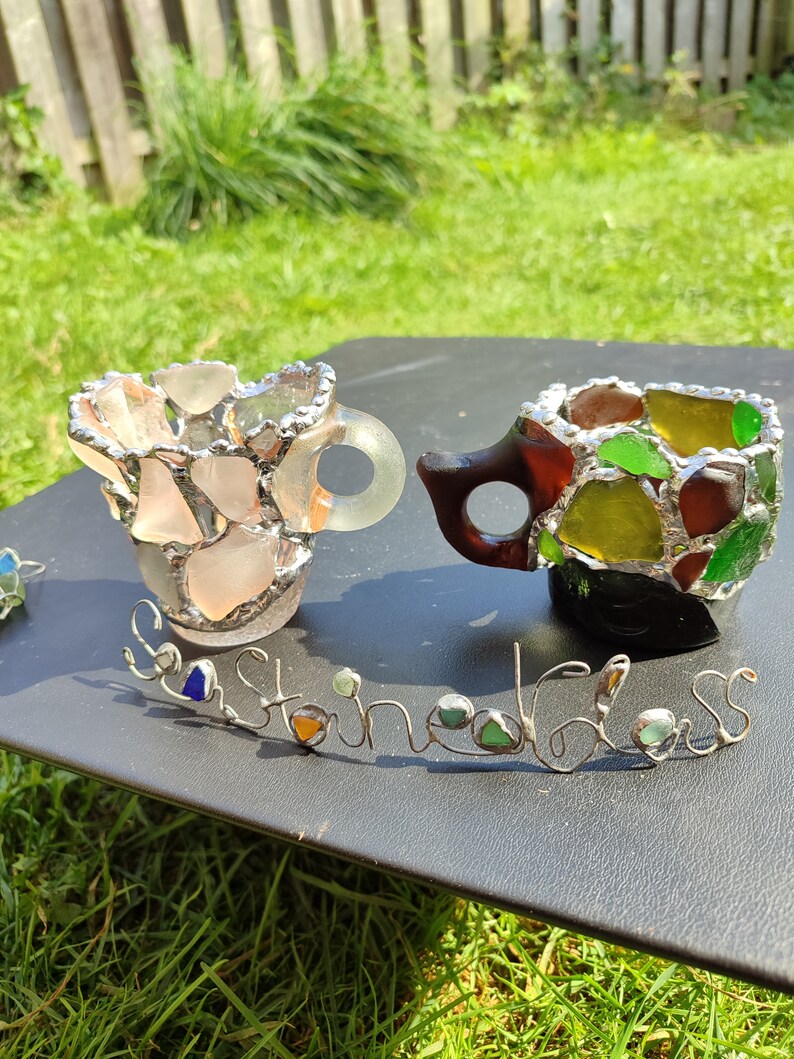 May include: Two small cups made of sea glass and wire. The cups are in the shape of a traditional teacup and are sitting on a black surface. The word "Sea Glass" is spelled out in wire with sea glass pieces used as the letters.