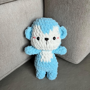 Puede incluir: Un muñeco de mono de ganchillo hecho a mano en tonos azul claro y blanco. El juguete de peluche tiene una cabeza redonda, orejas pequeñas y una cara sonriente con ojos negros y mejillas rosadas. El mono está sentado.