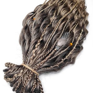 Puede incluir: Un conjunto de extensiones de cabello marrón y rubio con trenzas y plumas. Las extensiones están atadas juntas con una banda elástica.