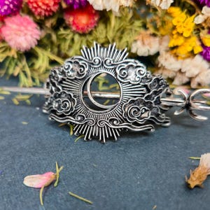 Op de afbeelding: Zilverkleurige haarspeld met een hemels ontwerp, met een maan, zonnestralen en wolken. De haarspeld heeft een lange pin om in het haar te bevestigen. De achtergrond bevat kleurrijke gedroogde bloemen.