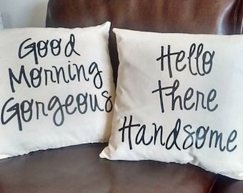 May include: Two white throw pillows with black lettering. One pillow says "Good Morning Gorgeous" and the other says "Hello There Handsome".
