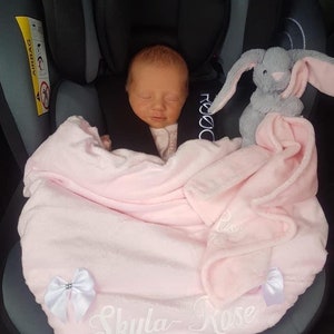 May include: A pink baby blanket with white embroidery that says "Skyla Rose". A gray bunny plush toy is on the blanket. A baby is sleeping in a car seat wrapped in the blanket.