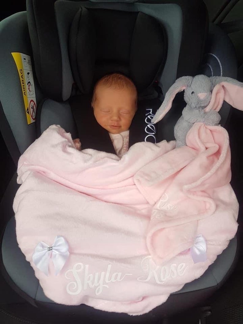 May include: A pink baby blanket with white embroidery that says "Skyla Rose". A gray bunny plush toy is on the blanket. A baby is sleeping in a car seat wrapped in the blanket.