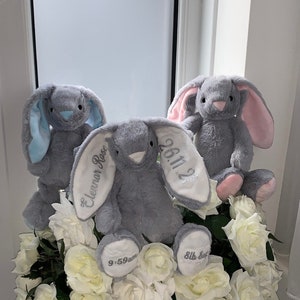 May include: Three gray plush bunny rabbits with embroidered details. The middle bunny has the text "Eleanor Rose" and "26.11.22" on its ears. The bunny on the left has blue ears and the bunny on the right has pink ears. All three bunnies are sitting in a bouquet of white roses.